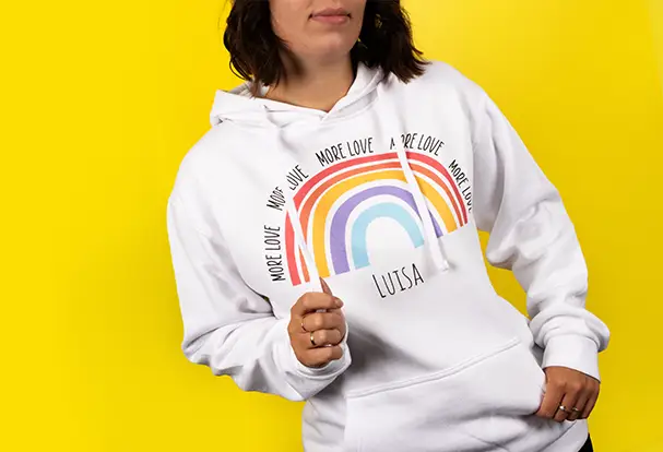 Neue Produkte: Hoodie für deine Lieblingsmotive Eine junge Frau im weißen Hoodie mit Regenbogen und ihrem Vornamen. Das neue Produkt steht im Fokus vor neutralem Hintergrund.