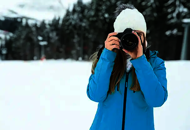 Fotografie-Tipps für beeindruckende Winterfotos Eine Frau in einer blauen Jacke fotografiert mit ihrer Kamera im Schnee. Entdecke jetzt unsere Foto Tricks für Winterfotos.
