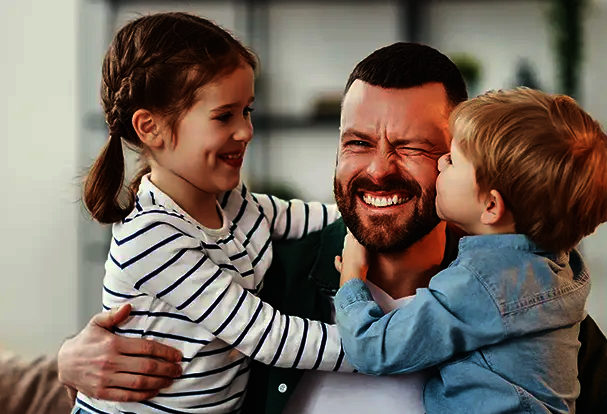 Fotografie Tipps – für emotionale Vatertagsfotos Strahlender Vater wird von seinen beiden Kindern umarmt und geküsst – Fotografie Tipps zum Vatertag.