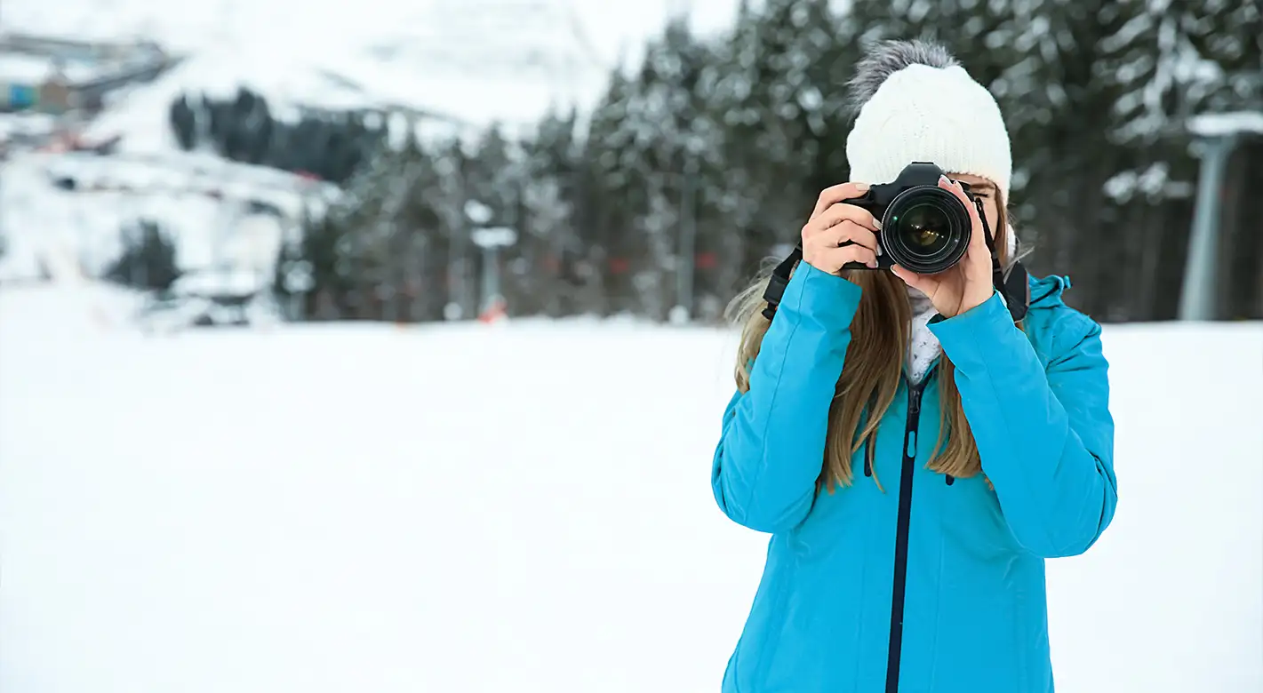 Eine Frau in blauer Winterjacke fotografiert im Schnee. Hol auch du dir Tipps für tolle Winterfotos!