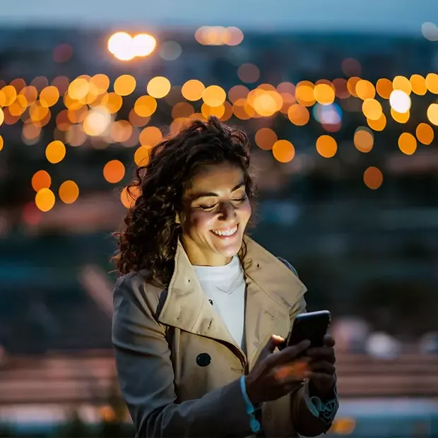 Abendstimmung: Ein Smartphone beleuchtet das Gesicht der im Vordergrund stehenden Frau. Dank des Bokeh-Effektes geben die Lichter der Stadt im Hintergrund einen wunderbaren Rahmen.