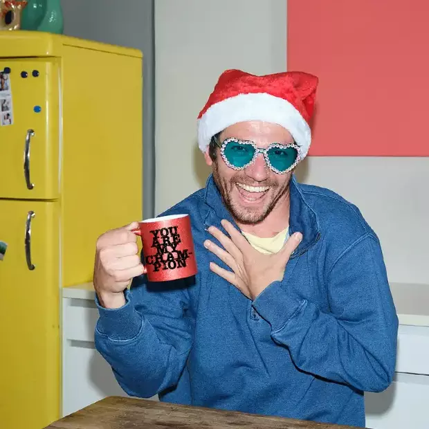 Festlicher Genuss mit Herz: Mann in Weihnachtsmütze und Herzchen-Brille, trinkt aus einer personalisierten Glitzertasse.