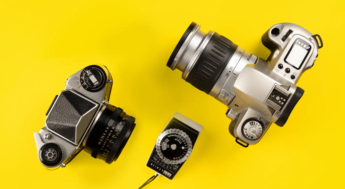 Zwei Vintage-Analogkameras in Schwarz und Silber liegen neben einem klassischen Belichtungsmesser auf einer gelben Fläche.