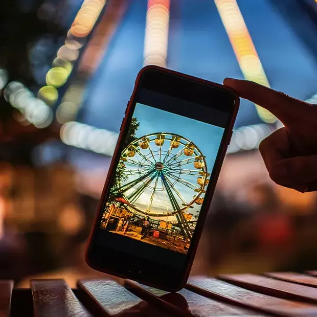 Riesenrad mal anders: Ein Smartphone im Vordergrund fängt das beleuchtete Riesenrad ein. Mit dem zweiten Handy wird ein Foto gemacht, welches das Riesenrad im Hintergrund durch einen Bokeh-Effekt magisch erscheinen lässt.