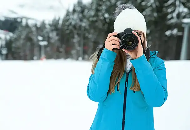 Eine Frau in einer blauen Jacke fotografiert mit ihrer Kamera im Schnee. Entdecke jetzt unsere Foto Tricks für Winterfotos.