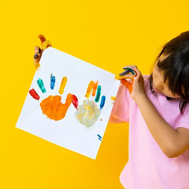 Ein Kind hält stolz sein selbst gestaltetes Bild in die Kamera – ein Kalender mit Kinderbildern. Auf weißem Papier sind bunte Handabdrücke zu sehen. Die Hände des Kindes sind noch voller Farbe.