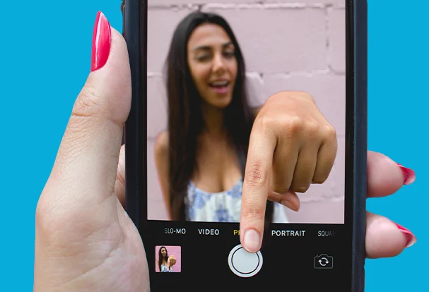 Foto-Tipps mit dem Smartphone oder der Kamera Eine junge Frau greift aus dem Smartphone heraus, um ein Foto von sich selbst zu machen. Unsere kreativen Foto-Tipps zeigen, wie man mit einfachen Mitteln beeindruckende Selbstporträts erzielen kann.