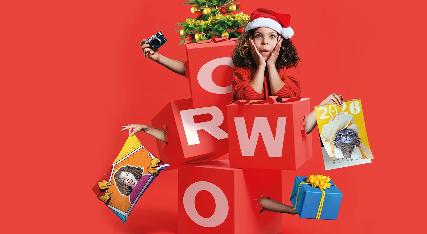 Fotoprodukte zu Weihnachten – kreative Geschenkideen von ORWO Kind in Weihnachtsoutfit überrascht zwischen großen ORWO-Geschenkboxen, hält verschiedene Fotoprodukte wie Fotokalender, personalisiertes Kissen, Geschenkbox und Kamera – Fotoprodukte zu Weihnachten bei ORWO.