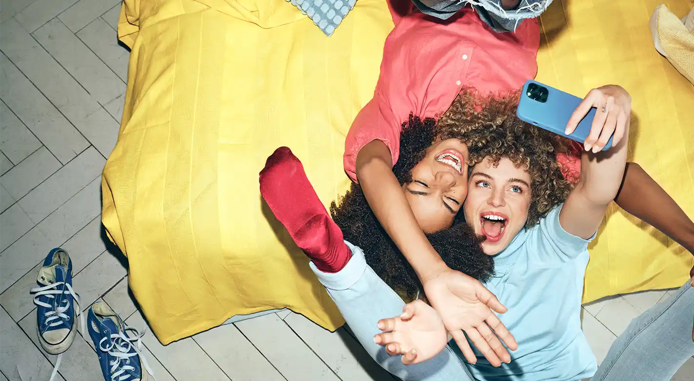 Spaß mit der Foto-App: Selfie-Momente festhalten Zwei junge Frauen machen mit ihrer Foto-App lustige Selfies, während sie auf einer Couch oder einem Bett liegen und ihre Füße in die Luft strecken.