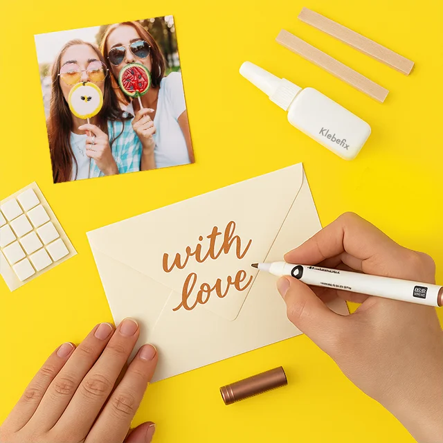 Fotozubehör und Kreativbedarf – Grußkarten und Fotogeschenke individuell gestalten Person schreibt „with love“ auf einen Briefumschlag, umgeben von Brushpen, Fotos und Klebepads.