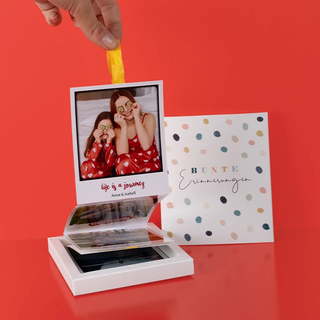 Kreatives Fotozubehör – stylische Box für Sofortbilder und besondere Erinnerungen Person zieht Sofortbilder mit gelbem Band aus einer Box, daneben liegt eine farbenfrohe Verpackung mit der Aufschrift „Bunte Erinnerungen“.