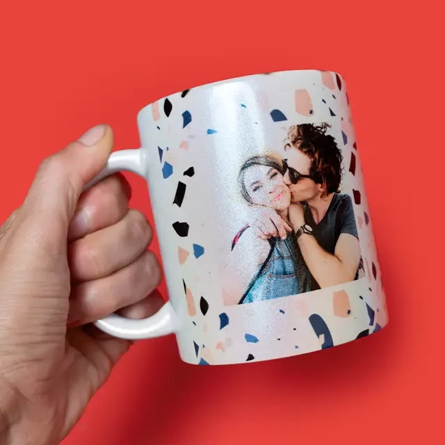 Eine Hand hält eine Tasse mit Perlmutteffekt vor einem roten Hintergrund. Die Tasse ist mit dem Foto eines Paares bedruckt.