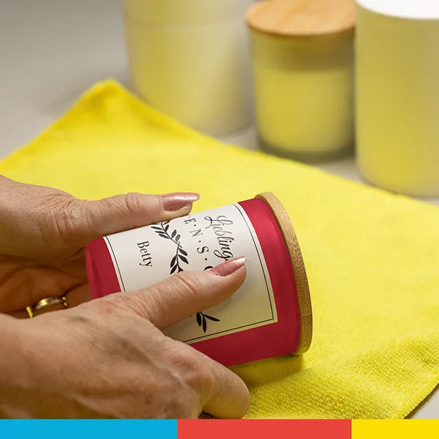 Kerze mit Foto, sorgfältig produziert Zwei Hände kleben sorgsam das personalisierte Etikett auf das Glas einer Kerze mit Foto auf.