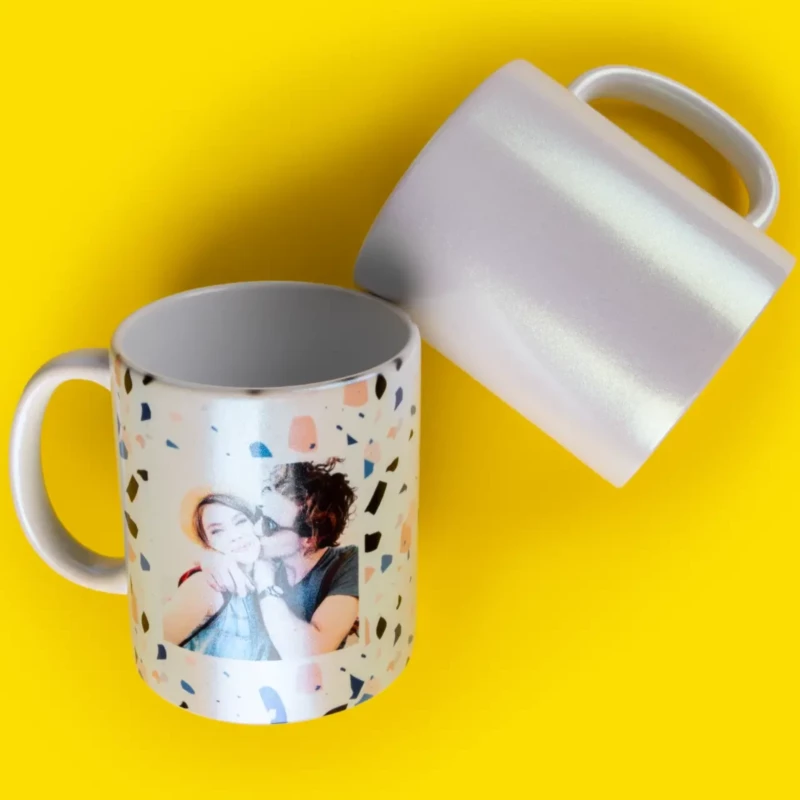 Zwei Perlmutt-Glitzertassen auf gelbem Hintergrund. Eine Tasse ist mit Foto gestaltet, die andere ist weiß.